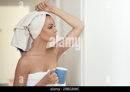 Pretty middle-aged woman wrapped in fresh clean white towels around her head and body leaning against an interior wall with a mug of beverage in a Stock Photo