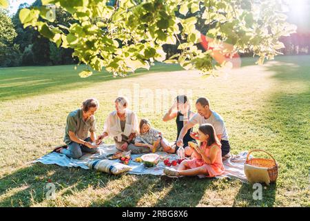 Big family under Linden tree on the picnic blanket on the in city park green grass. They are eating boiled corn, apples, peaches, pastries and waterme Stock Photo