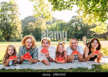 Big family lying on the picnic blanket in city park under Linden tree during weekend Sunday sunny day, smiling, laughing at camera and eating watermel Stock Photo