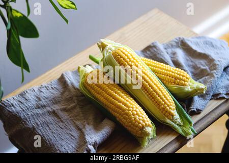 Three fresh corn cobs with leaves on a wooden table over grey wall background. Raw corncobs on light brown wooden table, rustic style. Corn on cobs li Stock Photo