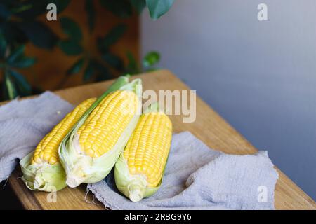 Stack of fresh corn cobs with leaves on a wooden table over grey wall background. Raw corncobs on light brown wooden table, rustic style. Corn on cobs Stock Photo