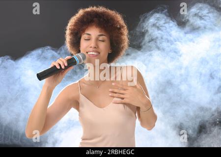 Curly young woman with microphone singing in smoke on black background Stock Photo