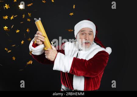 Emotional man in Santa Claus costume blowing up party popper on black background Stock Photo