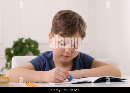 Little boy erasing mistake in his notebook at white desk indoors Stock Photo