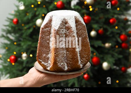 Woman holding delicious Pandoro cake decorated with powdered sugar near Christmas tree, closeup. Traditional Italian pastry Stock Photo