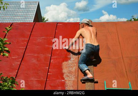 KYIV, UKRAINE - MAY, 15, 2023:  Roofer repainting old metal roof with a paint brush and red paint. Roofer painting house metal rooftop with red paint. Stock Photo