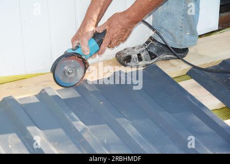 A building contractor is cutting lightweight metal, steel, corrugated, paint coated roofing sheet with an electric angle grinder laying profile sheet Stock Photo