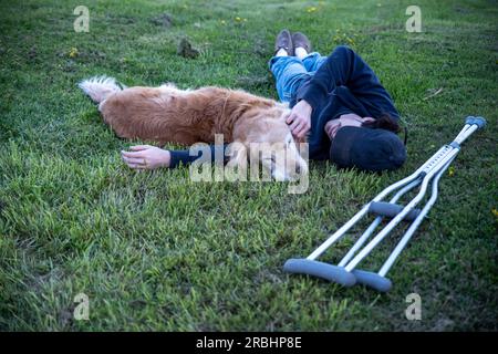 Crutches in the foreground of this idyllic rural nature background scene with a young man and his loving golden retriever dog laying on a grassy hill Stock Photo