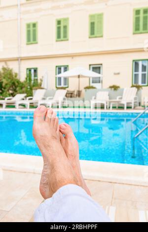 Barefoot male tourist relaxing by the outdoor swimming pool in seaside resort on sunny summer day, pov shot with selective focus Stock Photo