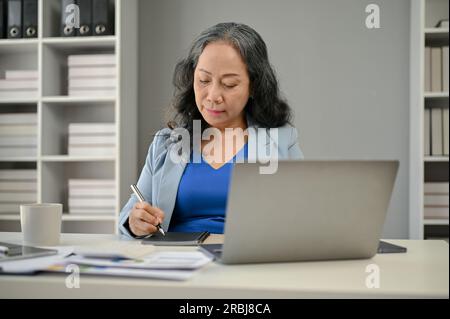 A professional Asian senior female boss focuses on her business work at her desk in the office. Businesspeople concept Stock Photo