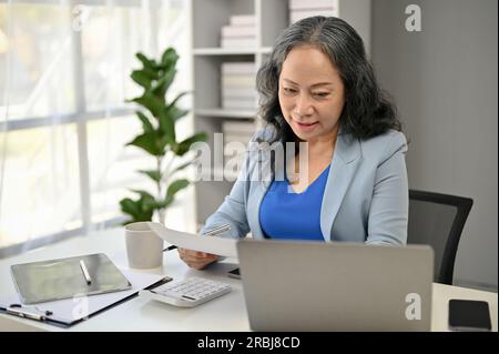 A professional and happy Asian senior female boss reading business documents and working on her tasks at her desk. Stock Photo