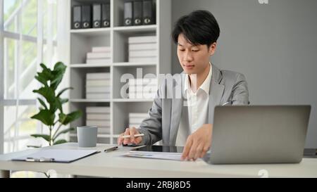 A professional and smart Asian businessman in a formal business suit focuses on reading business documents at his desk in the office. Stock Photo