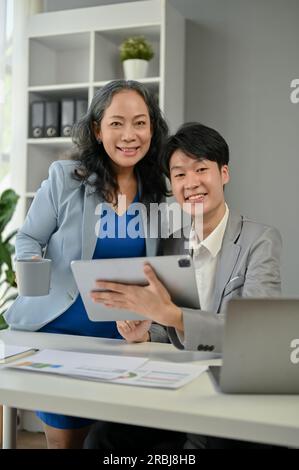 An attractive and professional mature Asian businesswoman and a young Asian businessman smiling and looking at the camera in the office. Stock Photo