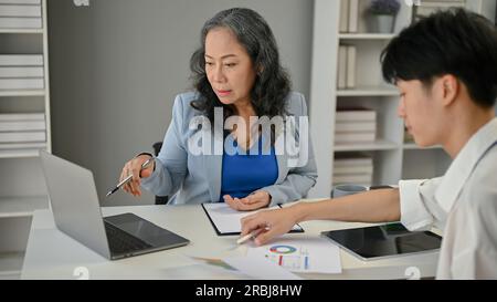 A professional and serious Asian senior businesswoman focuses on reading information on a laptop screen and working with a young male worker in her of Stock Photo
