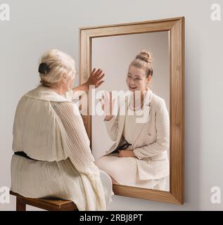 Creative conceptual collage. Tender image of senior woman looking in mirror and smiling to her young self reflection. Back to past Stock Photo