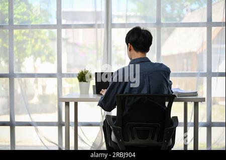 Back view image of a focused Asian businessman working on his work on his laptop computer at his desk by the window in a modern office. Stock Photo