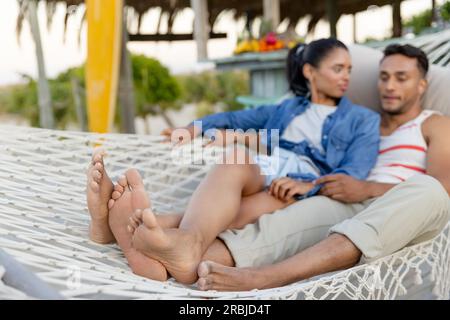 Biracial young couple with barefoot lying on hammock at beach during vacation. Unaltered, love, together, vacation, tourist resort, nature, relaxing, Stock Photo