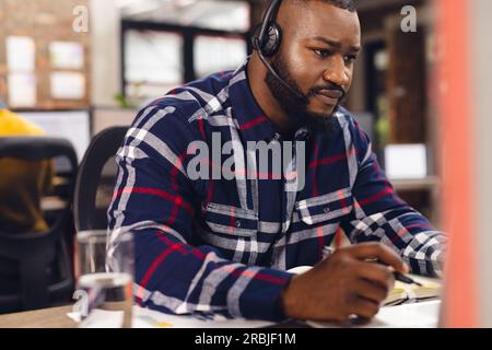 African american businessman sitting at desk and using phone headset at office. Casual business, communication and work, unaltered. Stock Photo