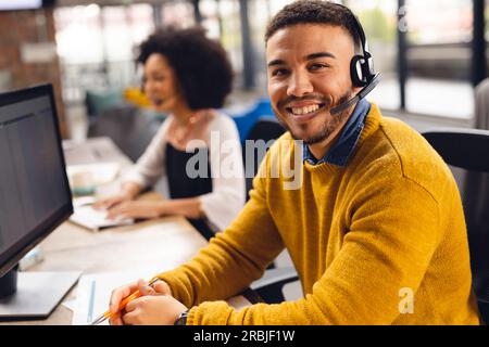 Portrait of happy biracial businessman using phone headset and smiling at office. Business, confidence, communication and work. Stock Photo