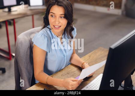 Happy caucasian businesswoman sitting at desk, using phone headset and computer at office. Casual business, communication and work, unaltered. Stock Photo