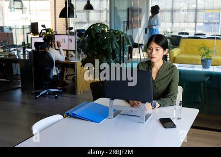 Focussed asian casual businesswoman sitting at desk using laptop on stand in office, copy space. Work, business, communication and casual office, unal Stock Photo