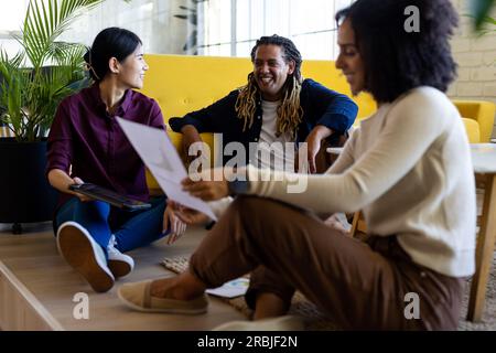 Three happy diverse casual business colleagues with tablet and document sitting on floor in office. Teamwork, work, business, meeting, communication a Stock Photo