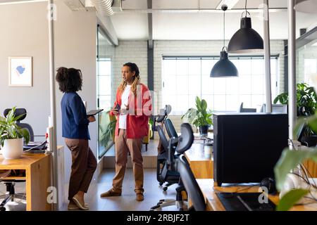 Two diverse casual business colleagues with tablet standing in office talking, copy space. Teamwork, work, business, communication, meeting and casual Stock Photo