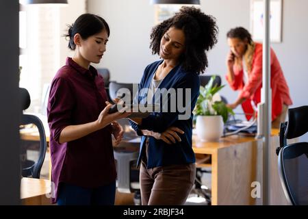 Two diverse female casual business colleagues using tablet standing in office talking. Teamwork, work, business, communication, meeting and casual off Stock Photo