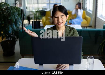 Happy asian casual businesswoman at desk in office having video call on laptop, smiling. Work, business, communication and casual office, unaltered. Stock Photo