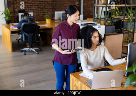 Two diverse female casual business colleagues using tablet and computer, discussing work at desk. Teamwork, work, business, communication and casual o Stock Photo