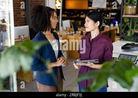 Two diverse female casual business colleagues with tablet standing in office talking. Teamwork, work, business, communication, meeting and casual offi Stock Photo
