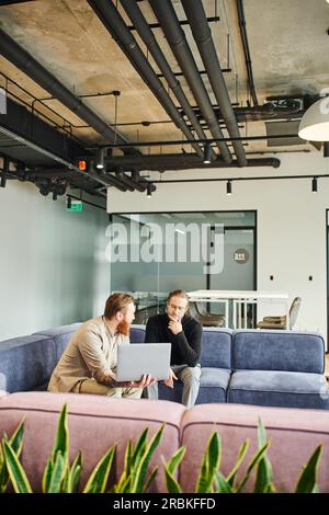 bearded businessman showing startup project on laptop to thoughtful colleague while talking on comfortable couch in lounge of modern office with high Stock Photo