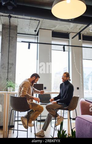 joyful and stylish entrepreneurs planning new business project while sitting near laptop in modern office environment with high tech interior, product Stock Photo