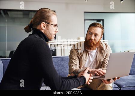 bearded businessman showing startup project on laptop to serious entrepreneur in black turtleneck and eyeglasses while sitting on couch in contemporar Stock Photo