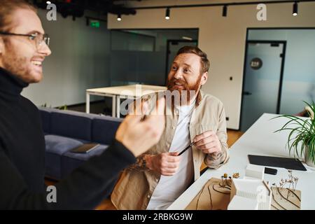 inspired architect in black turtleneck discussing innovative ideas with cheerful, bearded and tattooed colleague near building model in modern design Stock Photo