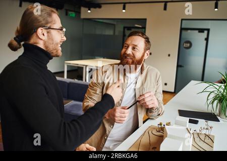 smiling bearded architect holding pen and looking at happy colleague talking near building model while discussing startup project in modern design stu Stock Photo