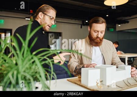 creative architect pointing at building model and discussing startup project with bearded colleague near blurred plant in contemporary design studio, Stock Photo