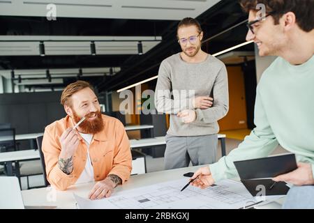 architect in eyeglasses holding notebook and pointing with pen at blueprint near cheerful, bearded and tattooed designer in contemporary coworking off Stock Photo