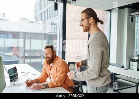 architect in eyeglasses smiling near excited bearded colleague sitting near laptop and blueprint while working on new startup project in contemporary Stock Photo