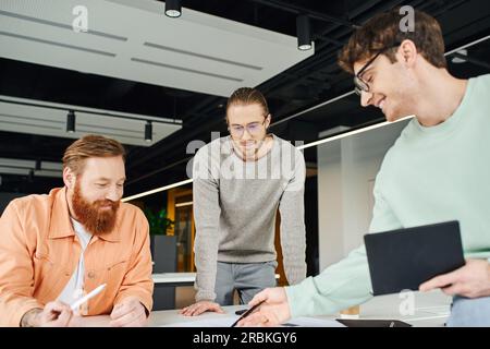 positive entrepreneur in eyeglasses holding notebook and pointing with pen near bearded architect and thoughtful colleague while working on business s Stock Photo