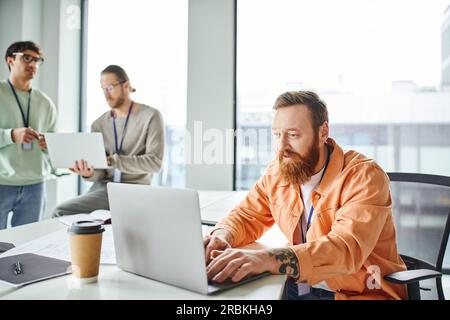 excited tattooed and bearded architect typing on laptop while working on new startup near takeaway drink, blueprint and colleagues on blurred backgrou Stock Photo
