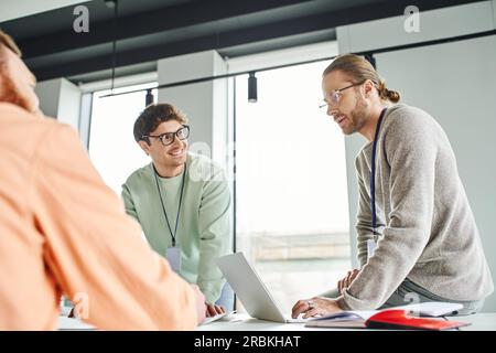 positive architectural designers in casual clothes looking at each other near laptop and businessman on blurred foreground while working on startup pr Stock Photo