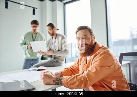 optimistic, tattooed and bearded designer looking at camera near blueprint and colleagues working on new business project on blurred background in con Stock Photo