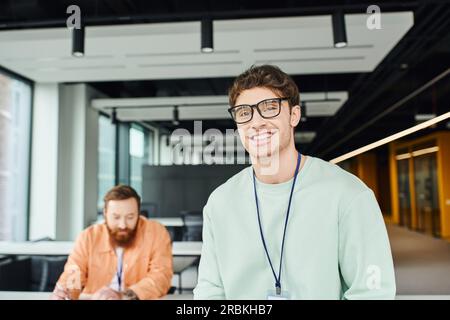 cheerful and optimistic architectural designer in eyeglasses smiling at camera near bearded colleague working on business startup on blurred backgroun Stock Photo