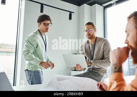 thoughtful architectural designer in eyeglasses working on laptop near thoughtful business colleagues and blueprint on blurred foreground, creative co Stock Photo