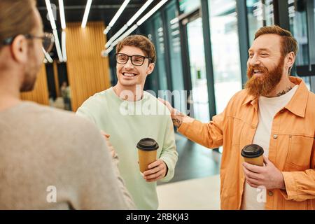 positive bearded man touching shoulder of happy colleague shaking hands with businessman on blurred foreground, successful entrepreneurs with coffee t Stock Photo