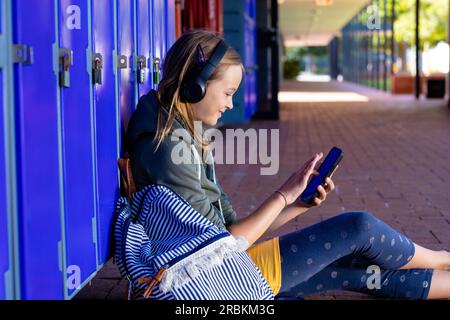 Smiling caucasian schoolgirl in headphones using smartphone sitting by lockers at school Stock Photo