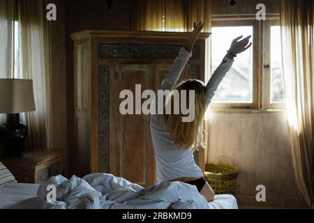 A young woman sits on the bed in the morning, stretches. Stock Photo