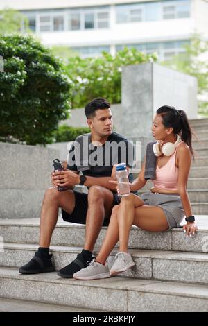 Fit couple sitting on steps, drinking water and talking after working out outdoors Stock Photo