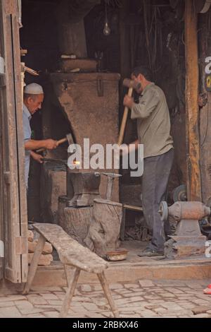 Two blacksmiths working in roadside workshop in Kashgar markets. One working at furnace and the other using sledgehammer. Clay furnace in centre. Stock Photo